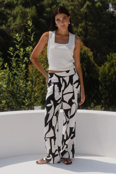 Woman wearing a white top and black and white patterned pants standing on a white architectural structure with greenery in the background.