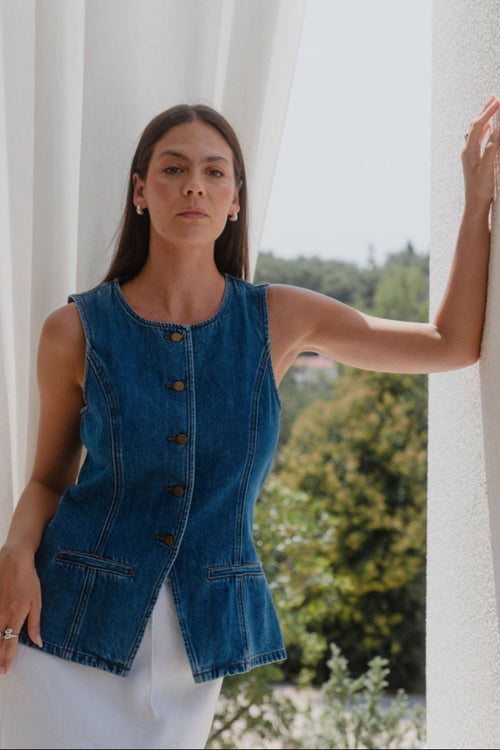 Woman wearing a denim vest standing against a white curtain with greenery in the background