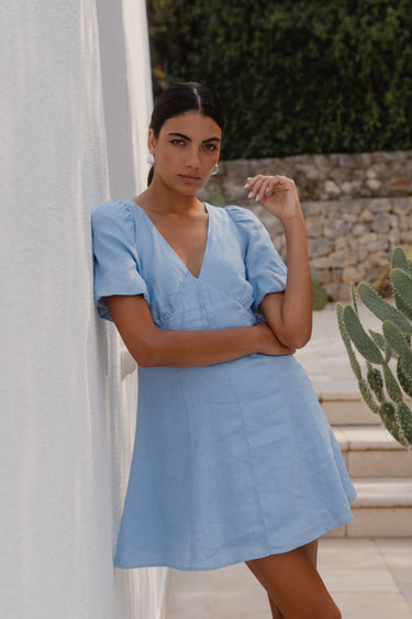 Woman in a light blue dress standing against a white wall with greenery in the background