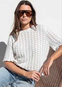 A woman wearing a white textured short-sleeve knit top and light blue jeans, sitting on a brown woven chair against a white wall.