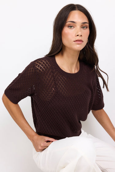 Woman wearing a dark brown knitted top and white pants against a white background