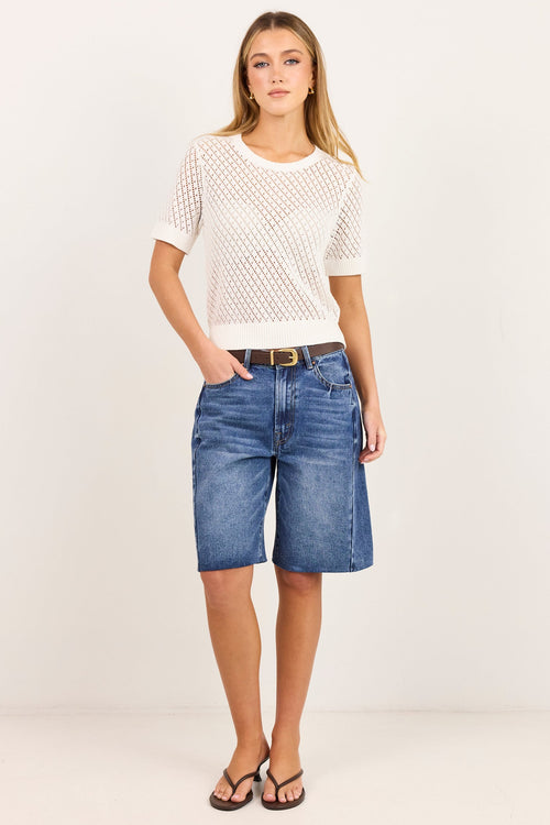 A woman modeling a white short-sleeve knit top and blue denim shorts against a plain white studio background.