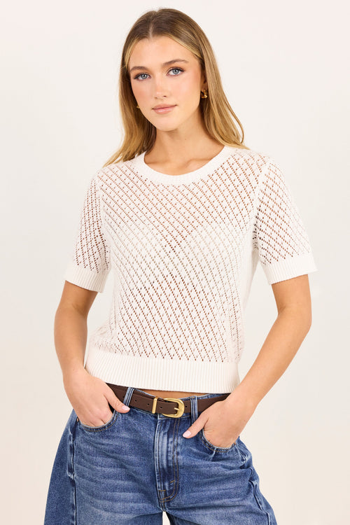 A woman modeling a short-sleeve knit top with a geometric openwork pattern in off-white and light brown, paired with blue jeans and a brown belt, against a plain light background.