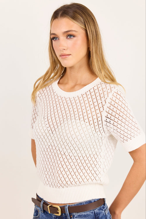 A woman wearing a short-sleeve knit top with a textured openwork pattern, paired with blue jeans and a brown belt, against a plain light background.
