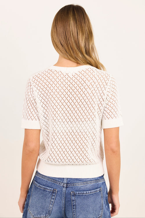 A woman viewed from the back wearing a short-sleeve knit top with a textured diamond pattern in off-white and light brown, paired with blue jeans, against a plain light background.