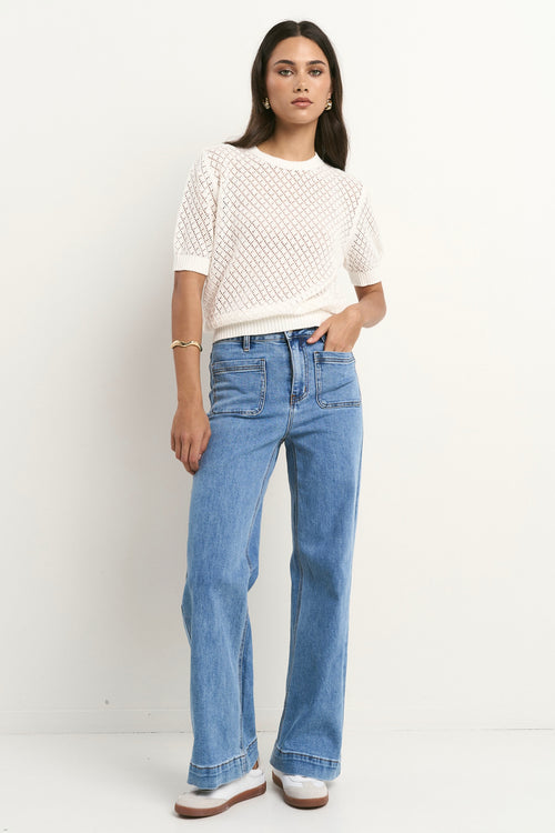 Woman wearing a short sleeve, open-knit ivory tee with a crew neck and light blue jeans.
