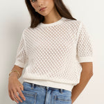 Woman wearing a short sleeve, open-knit ivory tee with a crew neck and light blue jeans.