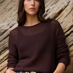A woman stands outdoors against a large rock formation, wearing a dark brown open-knit long-sleeve sweater, blue denim shorts, and a black belt with gold hardware.