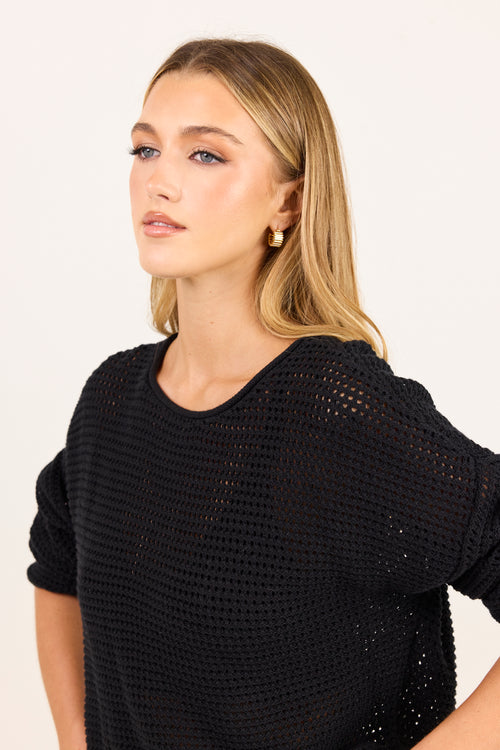A woman with blonde hair wearing a black open-knit short-sleeve top, photographed against a plain light background.