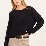 A woman modeling a black long-sleeve sheer knit top and beige straight-leg jeans with a black belt, standing against a plain off-white studio background.
