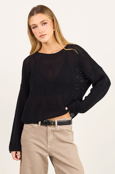 A woman modeling a black long-sleeve sheer knit top and beige straight-leg jeans with a black belt, standing against a plain off-white studio background.