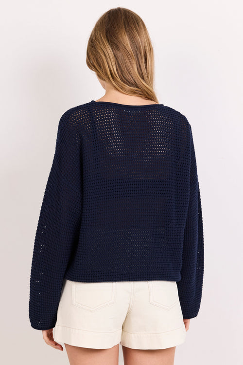 A woman viewed from the back wearing a dark navy open-knit cropped sweater and off-white shorts against a plain light background.