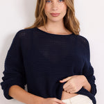 A woman wearing a dark navy blue open-knit long-sleeve sweater and off-white pants, standing against a plain white background.