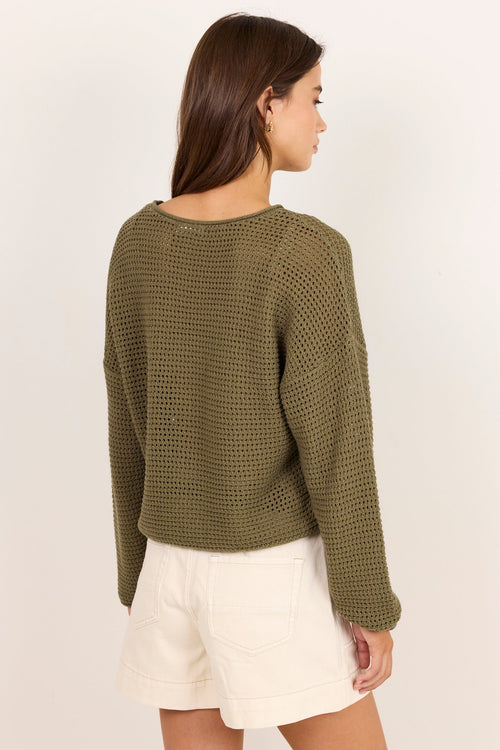 A woman shown from the back wearing an olive green textured knit long-sleeve sweater and off-white shorts, standing against a plain light beige studio background.