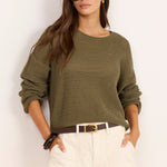 A woman modeling a textured olive green knit sweater and off-white shorts against a plain white background.
