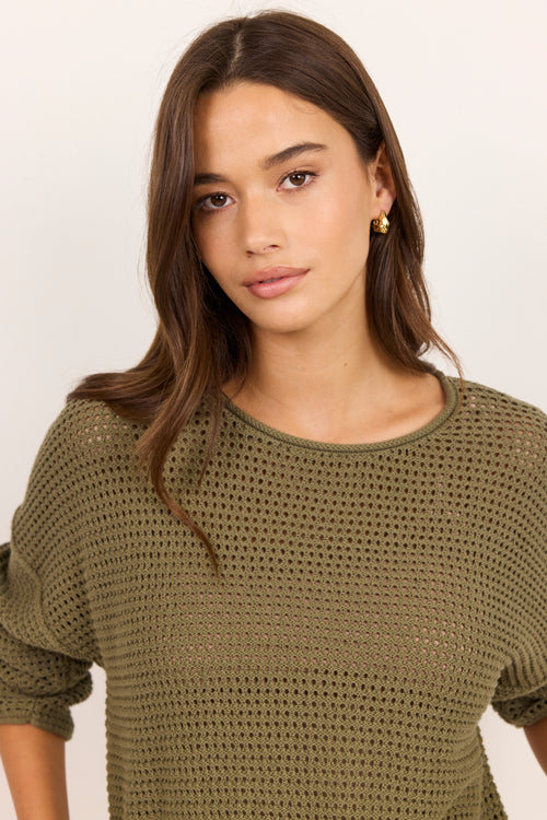 A woman wearing a short-sleeve olive green open-knit crewneck top, photographed against a clean off-white background.