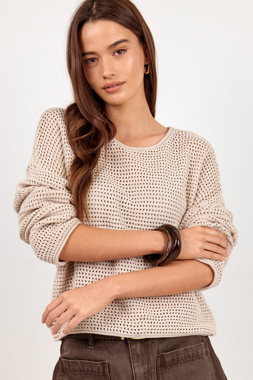 A woman modeling a beige open-knit long-sleeve sweater with dark brown pants against a plain light background.