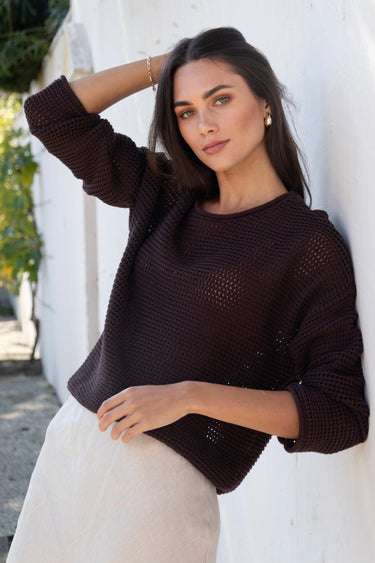 Woman wearing a dark brown knitted sweater against a white wall.