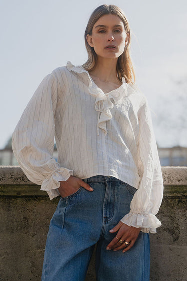 Woman wearing a white blouse with ruffles and blue jeans standing against a cityscape background.