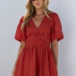 A woman wearing a rust-red short-sleeve mini dress and black sandals stands in a white-walled outdoor setting with a large white vase on the left.
