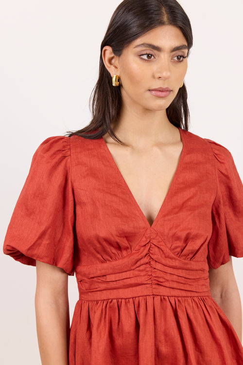 A woman wearing a rust-orange dress with puff sleeves, a deep V-neckline, and gathered ruching at the waist, photographed against a plain light background.