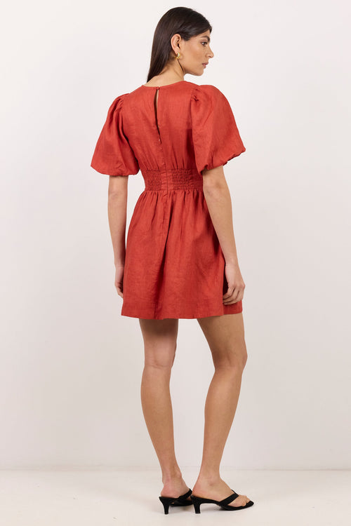 A woman modeling a rust-red mini dress from the back, featuring short puff sleeves, a gathered waist, and a back keyhole detail, paired with black mule sandals against a plain white studio background.
