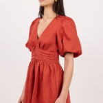 A woman modeling a terracotta orange short puff-sleeve dress with a gathered waist and deep V-neckline, photographed against a plain off-white studio background.