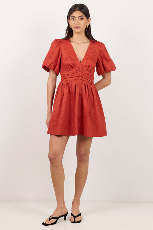 A woman modeling a terracotta-colored mini dress with puff sleeves, a deep V-neckline, and a gathered waist, paired with black sandals against a plain white studio background.