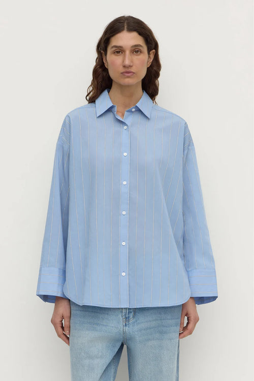 A woman wearing a light blue button-up shirt with thin vertical stripes and light-wash jeans, standing against a plain off-white background.