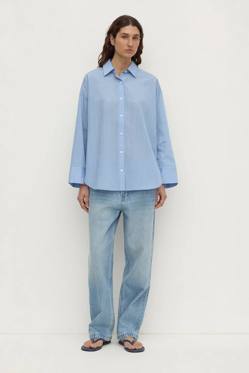 A woman stands against a plain off-white studio background wearing a light blue button-up shirt with subtle vertical stripes, light-wash blue jeans, and dark flip-flop sandals.