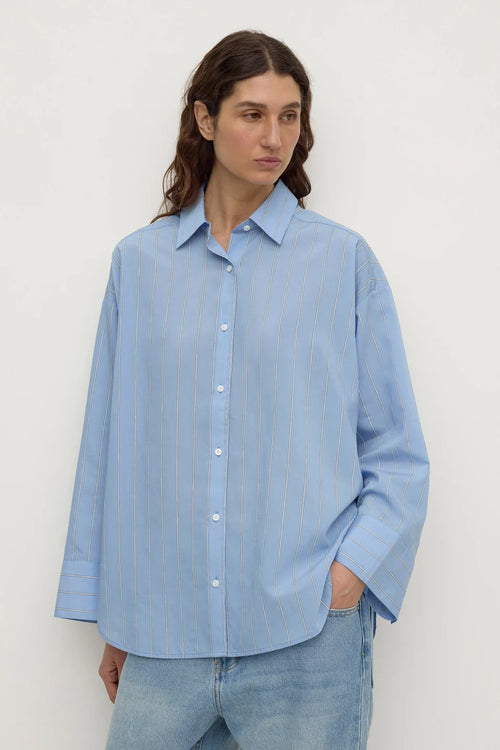 A woman wearing a light blue long-sleeve button-up shirt with subtle vertical stripes, paired with light-wash blue jeans, standing against a plain off-white background.