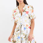 Woman wearing a cream floral mini dress with a tie belt, button front, short puff sleeves, and a drop waist design.