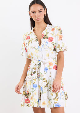 Woman wearing a cream floral mini dress with a tie belt, button front, short puff sleeves, and a drop waist design.