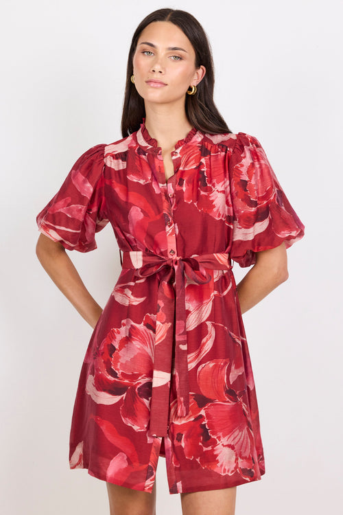 A woman modeling a red floral print mini dress with puff sleeves, a button-up front, and a matching waist tie, set against a plain light gray background.