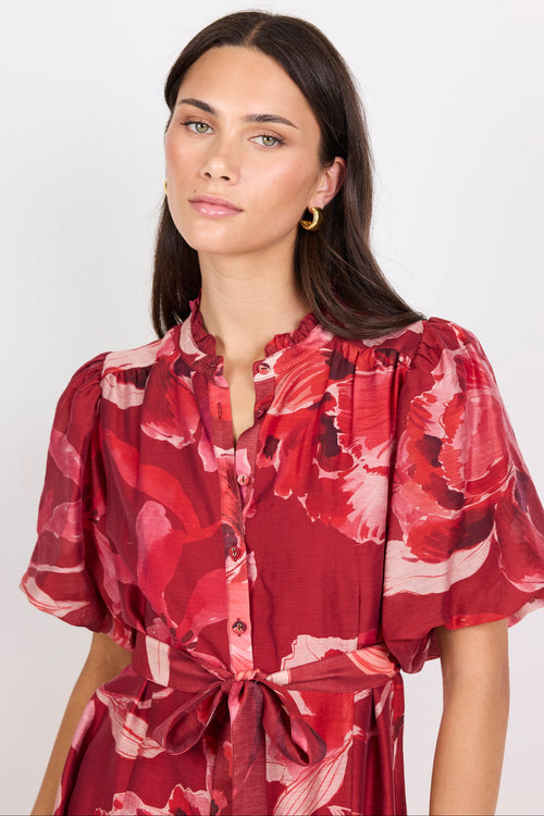 A woman wearing a red and pink floral print short-sleeve shirt dress with a tie waist, photographed against a plain light gray studio background.
