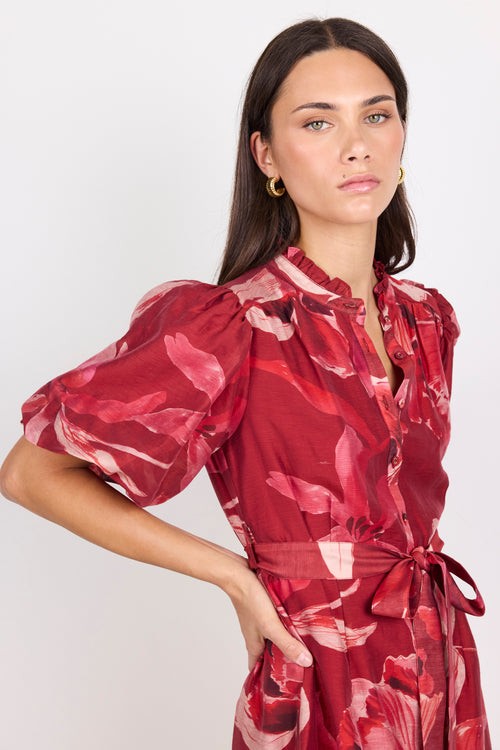 A woman modeling a red floral print dress with puff sleeves, a ruffled collar, and a matching waist tie, photographed against a plain light gray studio background.