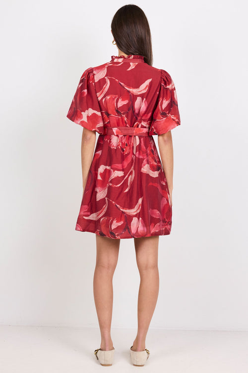 A woman viewed from behind wearing a short red dress with a pink and white floral pattern, standing against a plain white studio background.