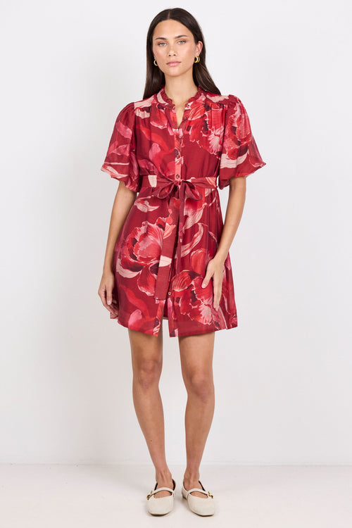 A woman modeling a short-sleeved red floral print dress with a tie waist, standing against a plain white studio background.
