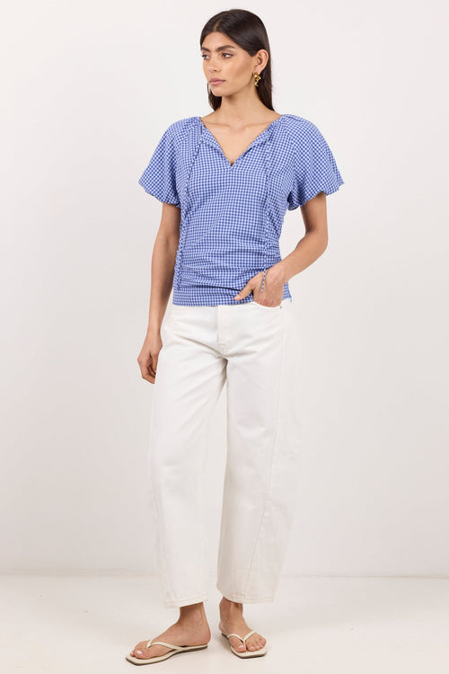 A woman modeling a blue and white gingham short-sleeve top with a split V-neck, paired with white cropped pants and flat sandals, standing against a plain white studio background.