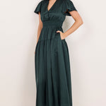 A woman modeling a dark green midi dress with short flutter sleeves, a V-neckline, and side pockets, standing against a plain off-white studio background.
