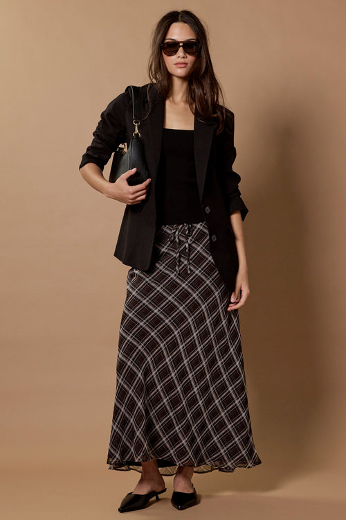 A woman models a black blazer, black top, and a black and white plaid maxi skirt against a solid beige studio backdrop.