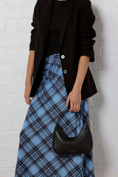 A model wearing a black blazer, black textured top, and a blue plaid skirt, holding a black crescent-shaped leather handbag against a white brick wall.