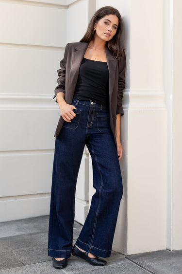 Woman in a black top and blue jeans leaning against a building wall.