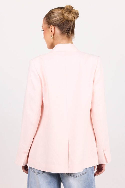 A woman viewed from behind wearing a light pink blazer and blue jeans against a plain white studio background.