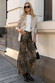 Woman in stylish outfit with leopard print pants and sunglasses standing against a building.