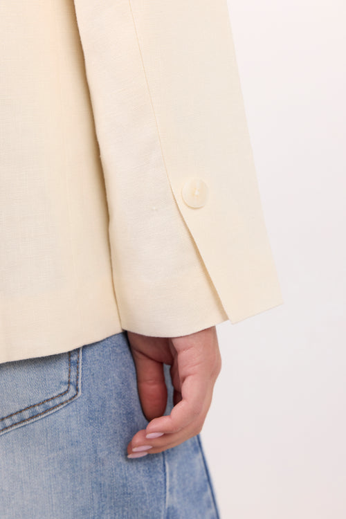 Close-up of a person wearing a light cream-colored blazer with a single visible button on the cuff, paired with light blue jeans against a plain light background.