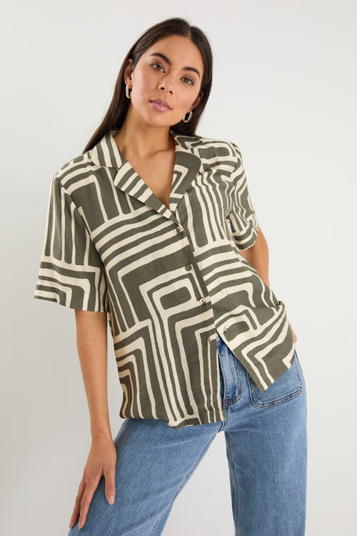 Woman wearing a short-sleeve shirt with an abstract olive and cream geometric print and button front, styled with mid-wash denim jeans.