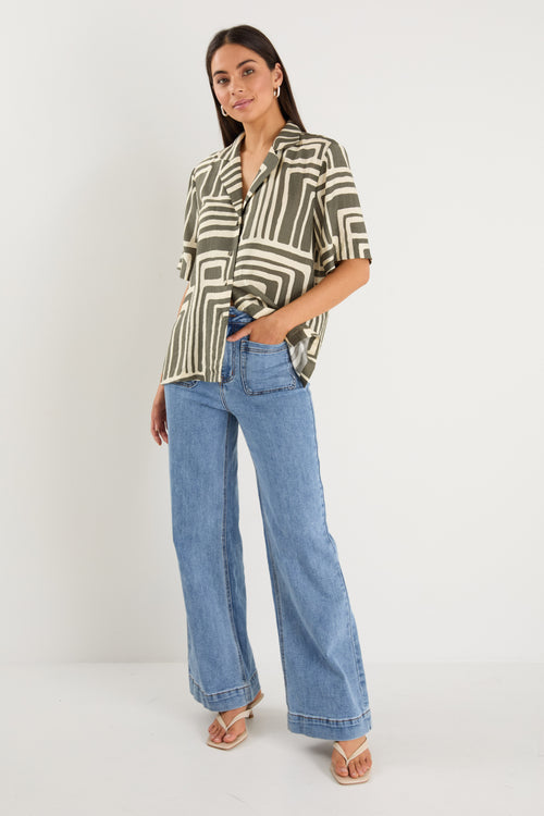Woman wearing a short-sleeve shirt with an abstract olive and cream geometric print and button front, styled with mid-wash denim jeans.