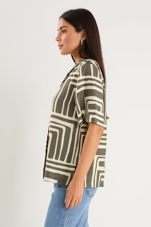 Woman wearing a short-sleeve shirt with an abstract olive and cream geometric print and button front, styled with mid-wash denim jeans.