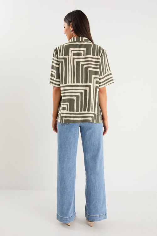 Woman wearing a short-sleeve shirt with an abstract olive and cream geometric print and button front, styled with mid-wash denim jeans.